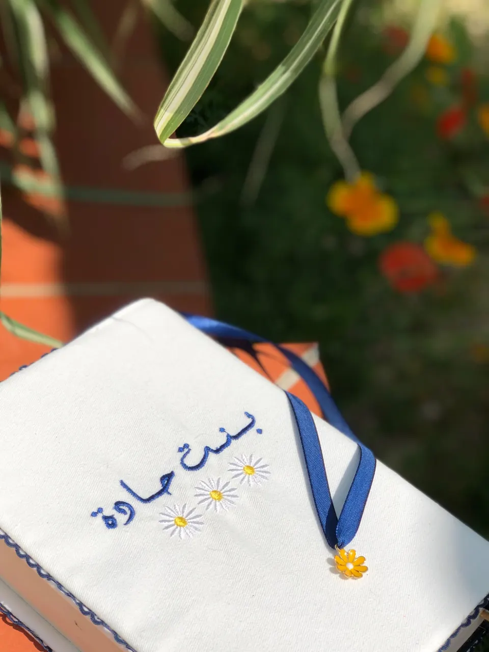 Protège-Coran en coton blanc cassé, texte arabe brodé bleu nuit et marguerites, ruban bleu et pendentif marguerite jaune
